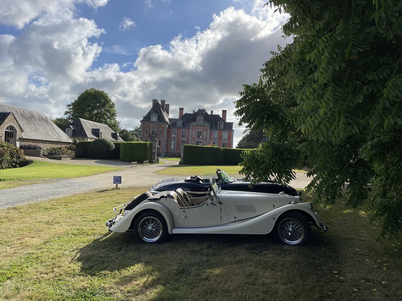 Zwei Oldtimer-Cabrios auf einer Rasenfläche vor einem kleinen Schloss mit Nebengebäuden und Kiesauffahrt.